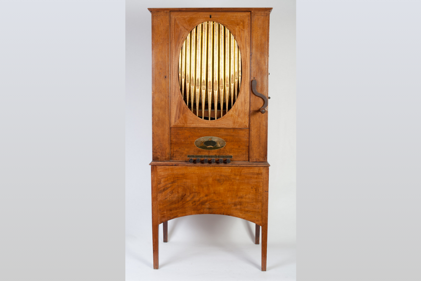 Hohe, hölzerne Salon-Orgel mit ovalem Messing-Pfeifenfeld und geschwungener Kurbel, freistehend auf vier Beinen.