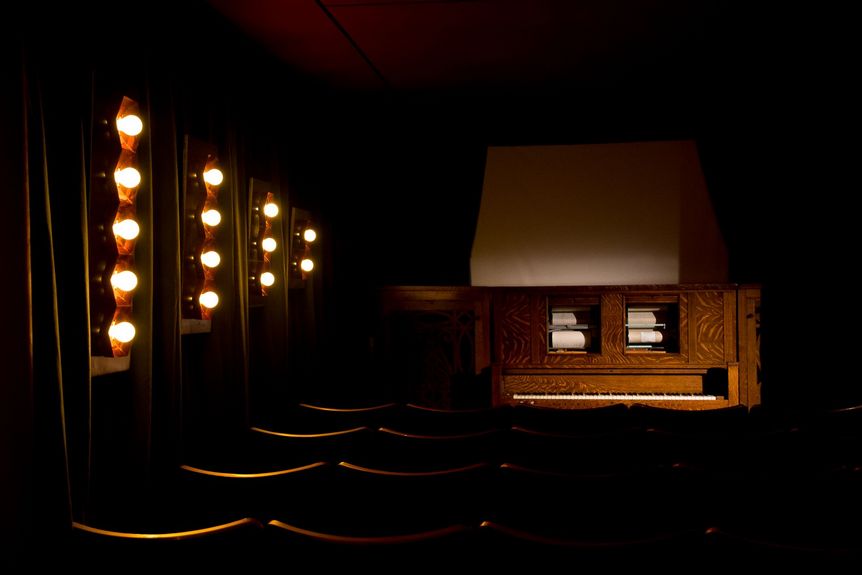 Dunkler Saal mit beleuchteten Wandleuchten, Blick auf eine große Kinoorgel mit Holzverkleidung vor Leinwand.